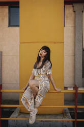 Full body of young self esteem ethnic female with long dark hair in stylish outfit sitting on metal railing near modern building and looking at camera - ADSF21098