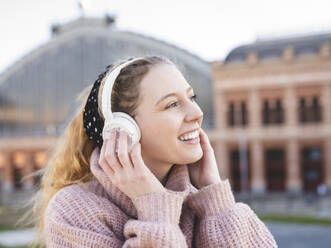 Seitenansicht einer fröhlichen Teenagerin mit langen gewellten Haaren in einem warmen Pullover, die lächelt, während sie mit Kopfhörern auf einem Stadtplatz Musik hört - ADSF21107