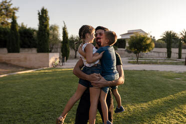 Father carrying daughter and son in arms while standing at park - AMPF00108