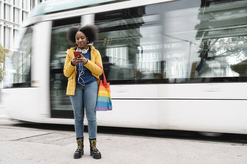 Young woman text messaging on mobile phone while standing at footpath against tram - JCZF00518