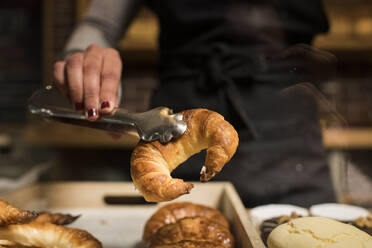 Young baker holding croissant in serving tongs at bakery - DAMF00735