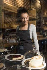 Lächelnde junge Bäckerin betrachtet einen Kuchen in einer Bäckerei - DAMF00736