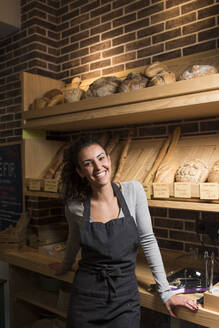 Lächelnder junger Besitzer steht vor einem Regal mit Backwaren in einer beleuchteten Bäckerei - DAMF00739