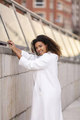 Lächelnde Frau mit Mantel, die sich auf eine Stützmauer am Fußweg stützt - MTBF00893