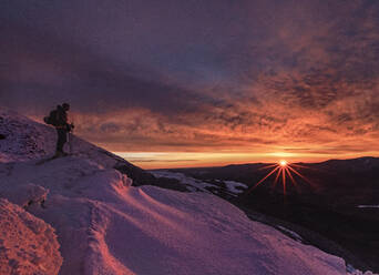 Skier stands on mountain in winter with stunning red pink sunrise - CAVF93715