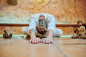 Anonymer flexibler Mann mit Tätowierung, der zwischen einer Buddha-Statuette und einer Gongschale an einer Steinmauer Yoga übt - ADSF21136