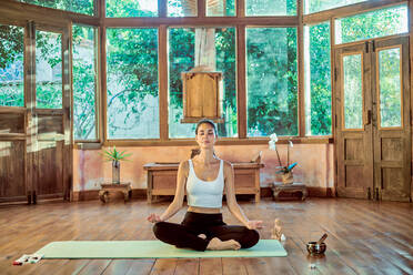 Junge konzentrierte Frau mit geschlossenen Augen praktiziert Yoga mit gekreuzten Beinen in der Nähe der Schale Gong im Haus - ADSF21146