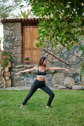 Side view of anonymous flexible female in sports clothes standing in Virabhadrasana pose on lawn in courtyard on summer day - ADSF21152