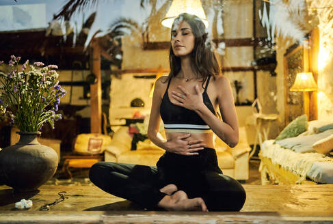Blick durch ein Fenster auf eine junge verträumte Frau mit geschlossenen Augen, die mit gekreuzten Beinen auf einer verglasten Terrasse sitzt und meditiert - ADSF21155