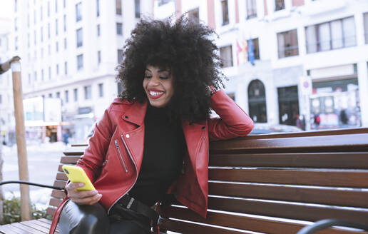 Stilvolle erstaunte afroamerikanische Frau, die auf einer Bank in der Stadt sitzt und Nachrichten auf ihrem Handy liest - ADSF21166