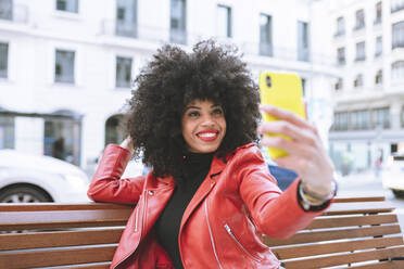 Selektiver Fokus einer fröhlichen Afroamerikanerin mit Afrofrisur, die auf einer Bank sitzt und ein Selbstporträt mit dem Handy aufnimmt - ADSF21172