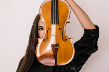 Female artist covering face with violin while standing against white background and looking at camera - ADSF21184