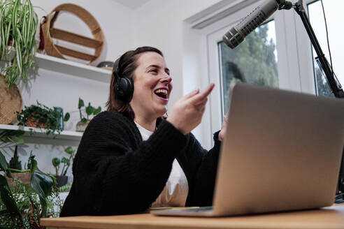 Laughing woman sitting at desk with laptop during podcast session at home - ASGF00031