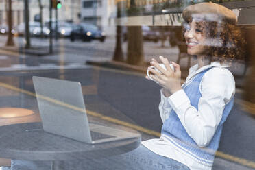 Smiling female teenager with laptop and coffee cup sitting by window at cafe - PNAF00923