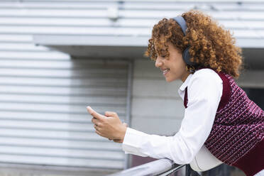 Smiling teenage girl with headphones using mobile phone while leaning on railing - PNAF00934