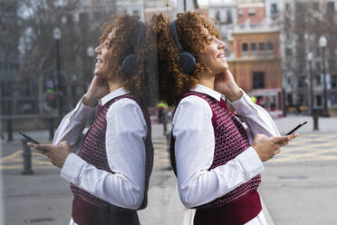 Fröhliche Frau mit Kopfhörern, die mit einem Mobiltelefon an einer Glaswand steht - PNAF00945
