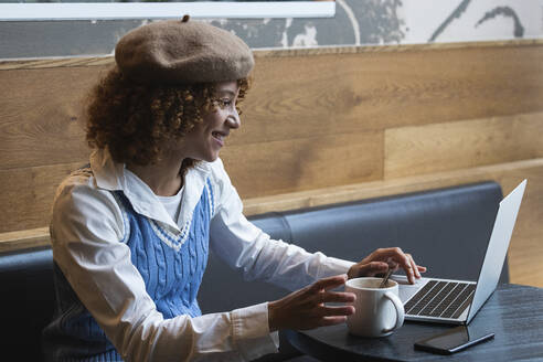 Lächelnder weiblicher Teenager mit Baskenmütze an einem Laptop in einem Café - PNAF00947