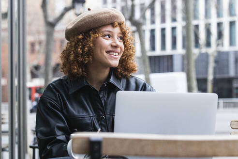Smiling teenage girl wearing beret sitting with laptop at sidewalk cafe - PNAF00952