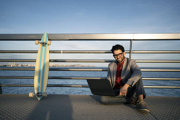 Smiling businessman with laptop sitting on pierbyt railing - RCPF00796