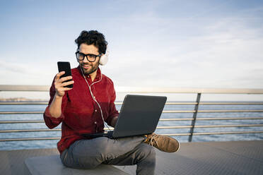 Smiling male entrepreneur with laptop listening music through headphones while using mobile phone on bench - RCPF00799