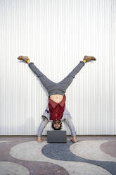 Businessman looking at laptop while doing handstand in front of wall - RCPF00802