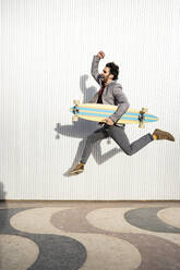 Fröhlicher Geschäftsmann mit Longboard springt auf dem Fußweg an der Mauer - RCPF00808