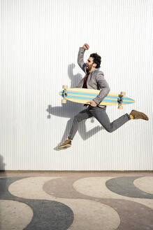 Cheerful businessman with longboard jumping on footpath by wall - RCPF00808