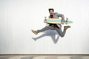Fröhlicher Mann hält Longboard und springt hoch vor die Wand - RCPF00809