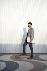 Smiling businessman with laptop standing on longboard by wall - RCPF00811