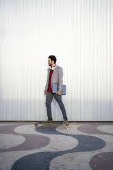 Male entrepreneur looking away while holding laptop on longboard in front of wall - RCPF00816