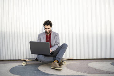 Smiling businessman with headphones using laptop while sitting on longboard in front of wall - RCPF00817