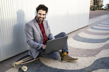 Lächelnder Geschäftsmann mit Laptop, der auf einem Longboard vor einer Wand sitzt - RCPF00818