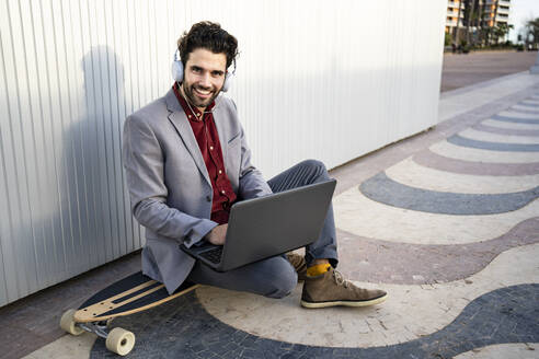 Lächelnder Geschäftsmann mit Laptop, der auf einem Longboard vor einer Wand sitzt - RCPF00818