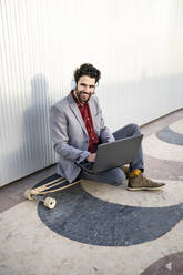 Smiling male entrepreneur with laptop sitting on longboard in front of wall - RCPF00819