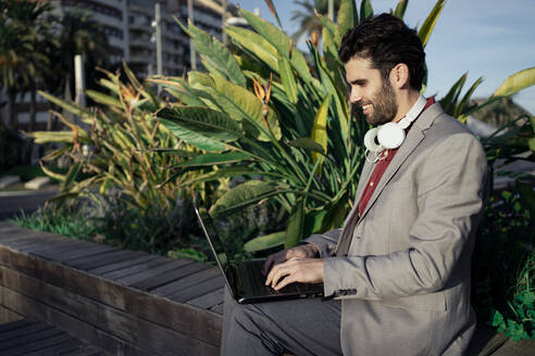 Smiling businessman using laptop while sitting on retaining wall - RCPF00820
