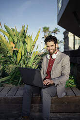 Lächelnder männlicher Geschäftsmann mit Laptop, der auf einer Stützmauer sitzt - RCPF00824