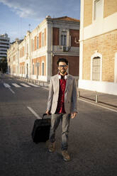 Smiling male entrepreneur with suitcase walking on road - RCPF00830