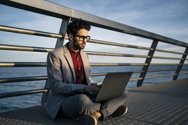 Männlicher Geschäftsmann, der einen Laptop benutzt, während er im Schneidersitz auf einem Steg sitzt - RCPF00833