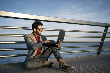 Smiling businessman using laptop while sitting on pier in front of railing - RCPF00835