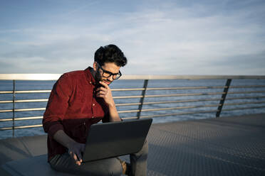 Nachdenklicher Geschäftsmann schaut auf seinen Laptop, während er auf einer Bank am Meer sitzt - RCPF00838