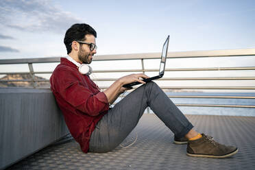 Smiling businessman using laptop on pier leaning against bench - RCPF00840