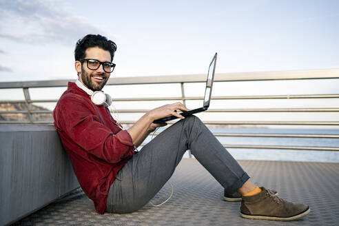 Lächelnder männlicher Geschäftsmann mit Laptop, der auf einem Steg sitzt und sich gegen eine Bank lehnt - RCPF00841
