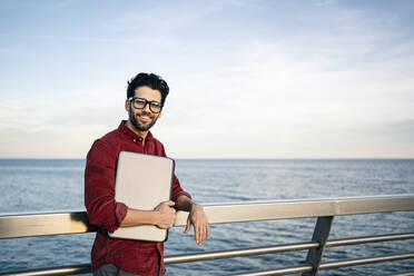 Smiling businessman with laptop leaning on railing by the sea - RCPF00842