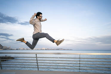 Carefree male entrepreneur with arms raised jumping by railing in front of blue sky - RCPF00849