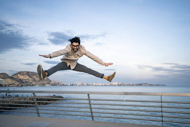Male business professional with arms outstretched doing splits in mid-air in front of blue sky - RCPF00850