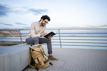 Lächelnder männlicher Unternehmer, der auf einer Bank am Pier sitzt und Tagebuch liest - RCPF00852