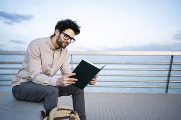 Lächelnder Geschäftsmann liest Tagebuch, während er auf einer Bank vor dem blauen Himmel sitzt - RCPF00854