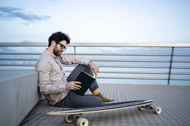 Businessman reading diary while sitting by longboard on pier - RCPF00855