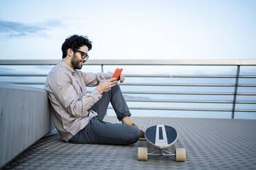 Lächelnder Geschäftsmann, der ein Mobiltelefon auf einem Longboard benutzt, während er auf einem Pier sitzt und sich gegen eine Bank lehnt - RCPF00857