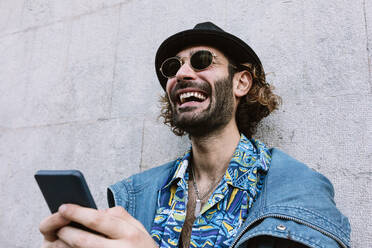 Lachender gut aussehender Mann mit Sonnenbrille, der sein Smartphone vor einer Wand hält - XLGF01285